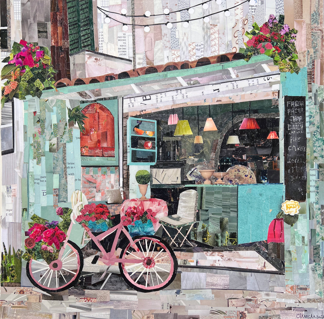 Chrissy McIntyre, Agios Gordios, paper collage on board, 14 x 14 inches. A pink bicycle with flower-filled baskets parked in front of an open café with mint-green walls, a terracotta tile roof, colored pendant lamps, a chalkboard menu, a white folding chair, and string lights overhead.