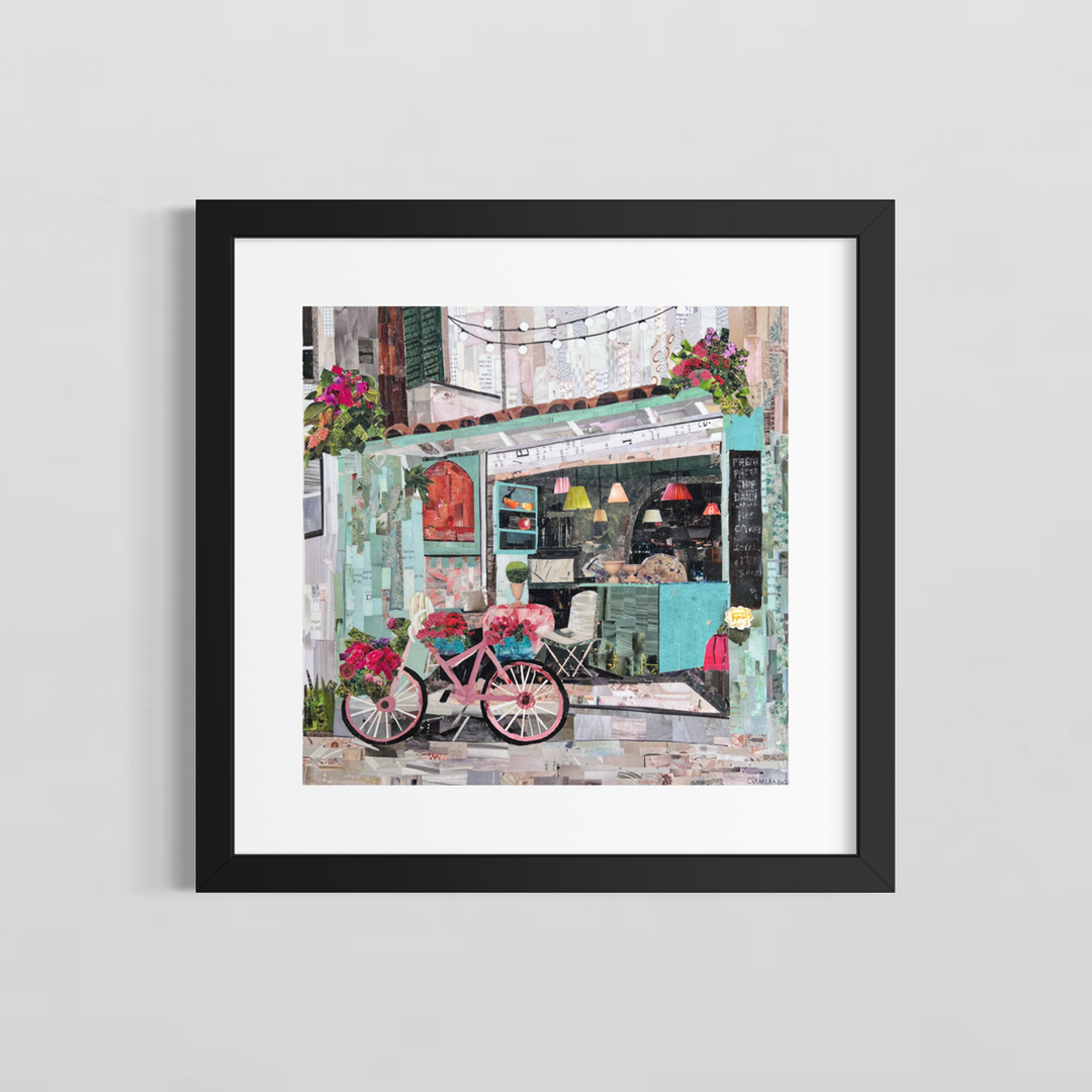 Chrissy McIntyre, Agios Gordios, paper collage on board, 14 x 14 inches, shown in a black frame with white matting on a light grey wall. A pink bicycle with flower baskets parked in front of a mint-green café with pendant lamps and a terracotta tile roof.