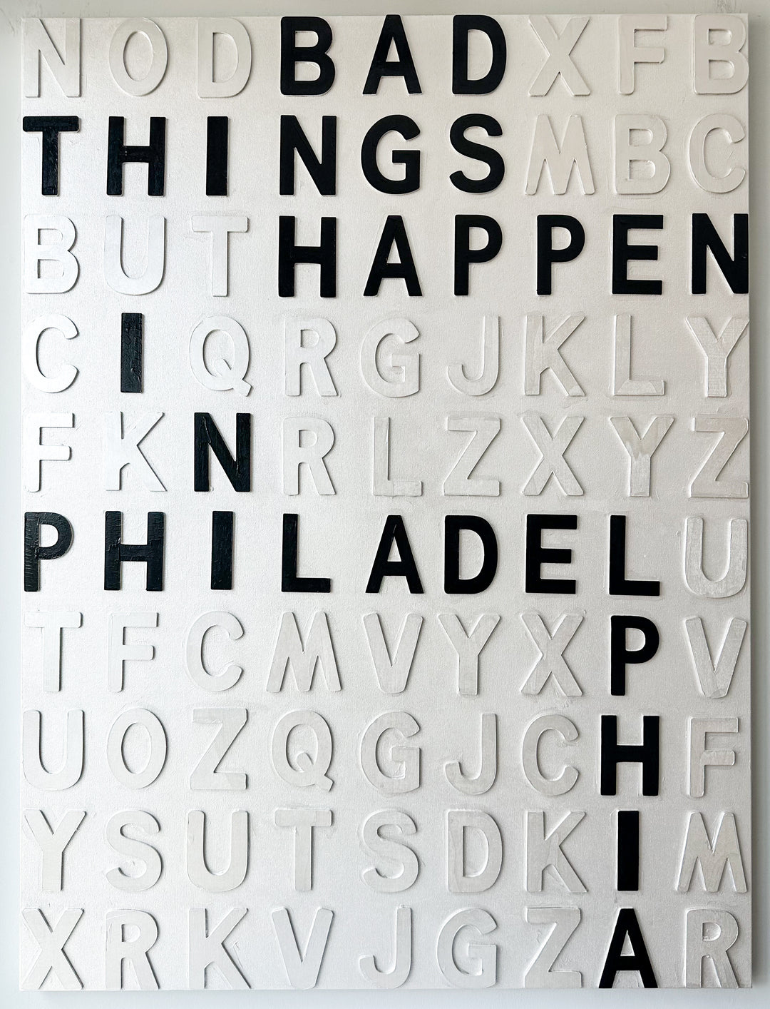 Steffanie Gerkin’s Bad Things White 3D is a contemporary mixed-media typography artwork made with raised wood and acrylic letters on canvas, featuring a white alphabet grid with bold black letters that spell “Bad Things Happen in Philadelphia.”