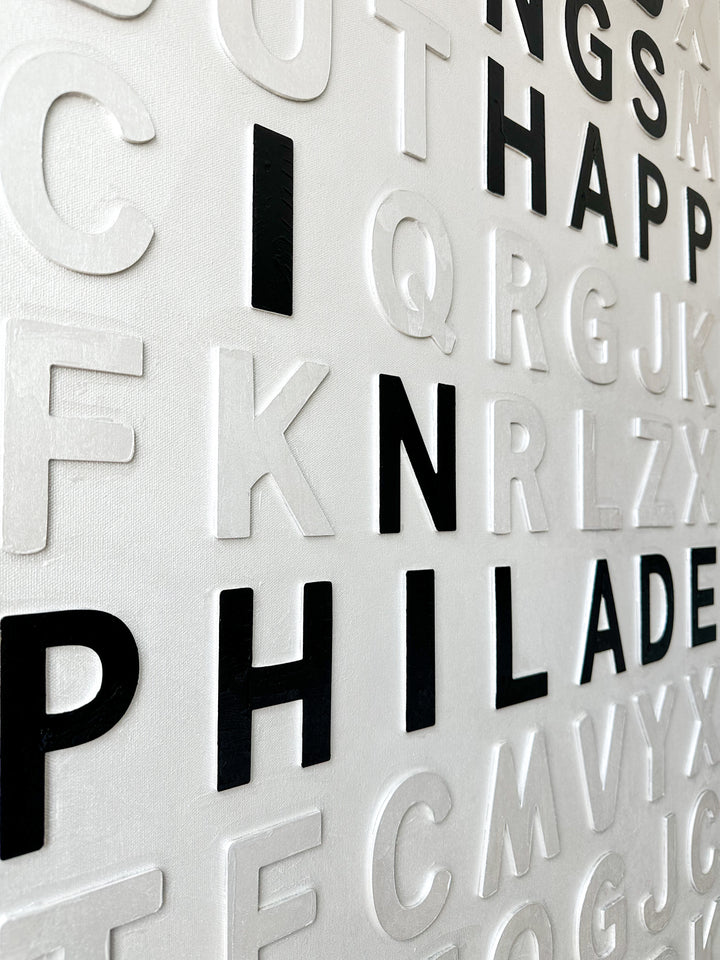 Steffanie Gerkin’s Bad Things White 3D is a contemporary mixed-media typography artwork made with raised wood and acrylic letters on canvas, featuring a white alphabet grid with bold black letters that spell “Bad Things Happen in Philadelphia.”