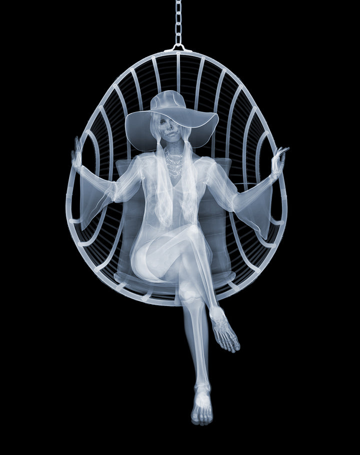 Framed Chromaluxe black and white x-ray photograph by Nick Veasey of a skeleton woman sitting in a swing chair with long pigtails, large hat and boho style dress crossing her legs
