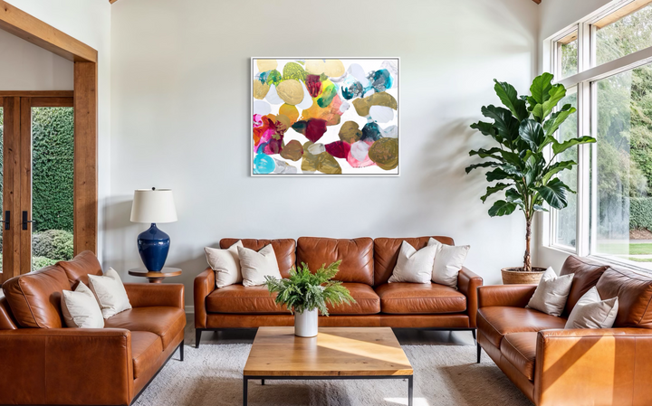 Liz Barber, Butterflies 3, acrylic paint, ink, and watercolor on canvas, 60 x 48 inches. Abstract composition of rounded shapes in metallic gold, olive, crimson, magenta, orange, yellow, turquoise, teal, and white on a white background, shown in a white frame in a living room with cognac leather sofas, a wood coffee table, and a fiddle-leaf fig by large windows.