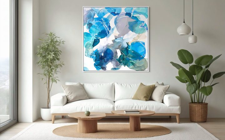 Liz Barber, Dancing Wave 3, acrylic paint, ink, watercolor, graphite, and pastel on canvas, 60 x 60 inches. Square abstract composition of overlapping translucent shapes in cobalt, cerulean, turquoise, teal, lavender gray, and pale beige with white linear marks, shown in a white frame above a white sofa with round wood coffee tables and potted plants beside a large window.