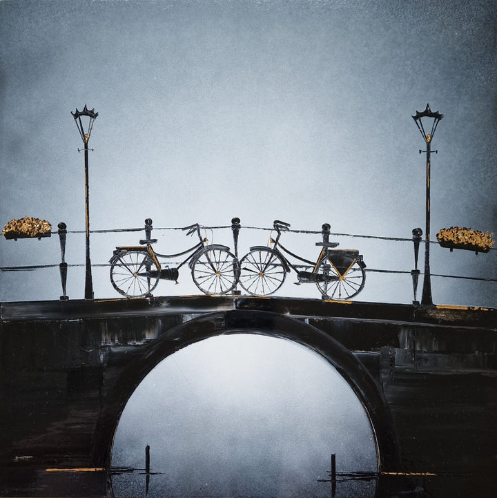 "Enjoy the Moment" by French artist Julien Rey — a mixed-media painting on board of two bicycles on a Parisian stone bridge, crafted with Chinese lacquer and gold leaf. Two classic city bikes with gold leaf accents rest against an iron railing flanked by tall street lamps and gold leaf flower baskets, above a luminous arched bridge reflection in misty water below. Original contemporary romantic Parisian bicycle painting on board available at Morton Contemporary gallery.