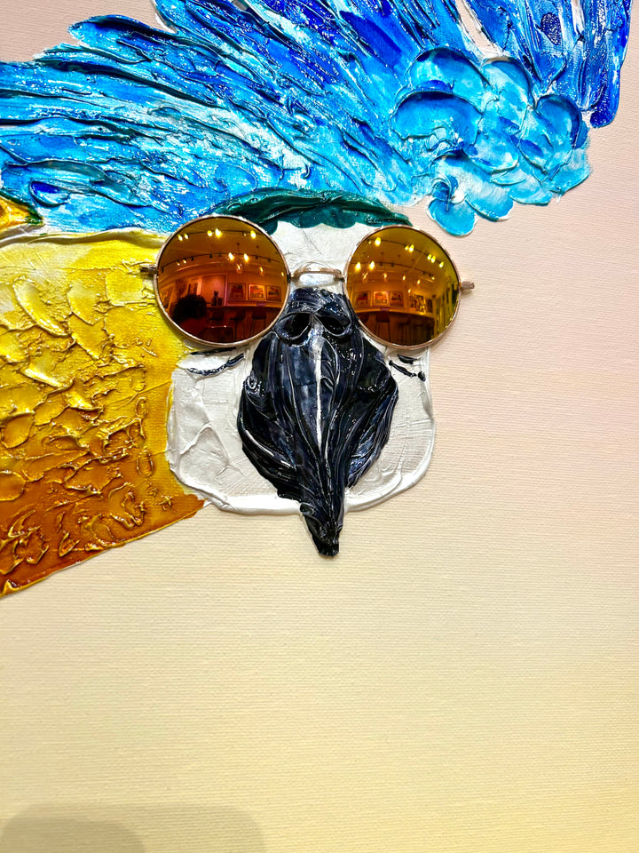 30” x 30” painting by French artist Moogly of a blue and yellow parrot in sunglasses with a pink and yellow ombre background. The parrot is depicted in mid-flight and made out of resin, giving it a textured and shiny finish. The sunglasses are real round, reflective sunglasses that the artist has adhered to the canvas.