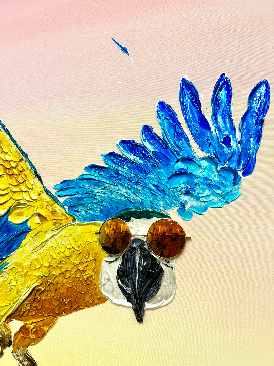 30” x 30” painting by French artist Moogly of a blue and yellow parrot in sunglasses with a pink and yellow ombre background. The parrot is depicted in mid-flight and made out of resin, giving it a textured and shiny finish. The sunglasses are real round, reflective sunglasses that the artist has adhered to the canvas.