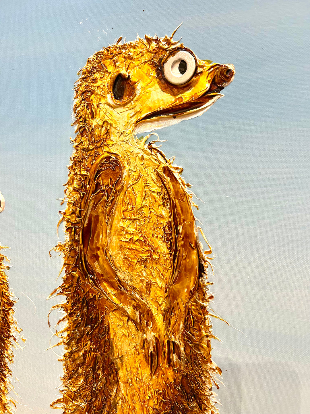 30” x 30” painting by French artist Moogly of three goldish-brown merecats. The two merecats on the sides look left and right while the smallest one in the middle looks straightforward with a light blue background. The merecats are made out of resin, giving them a textured and shiny finish. Their eyes are made out of washers that the artist has adhered to the canvas.