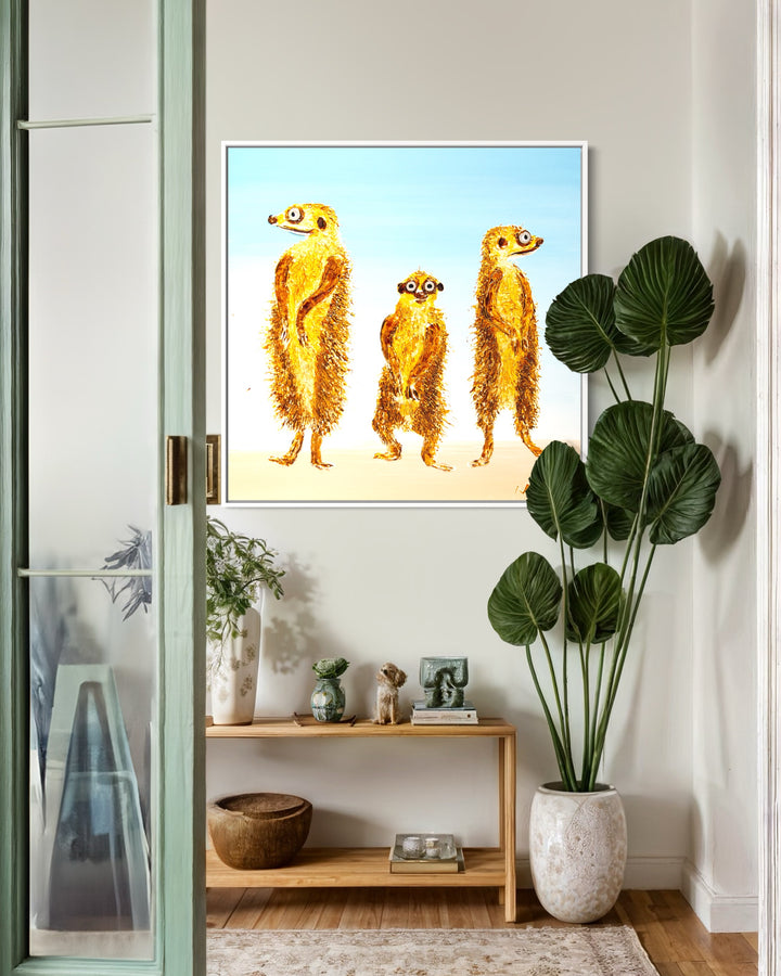 30” x 30” painting by French artist Moogly of three goldish-brown merecats. The two merecats on the sides look left and right while the smallest one in the middle looks straightforward with a light blue background. The merecats are made out of resin, giving them a textured and shiny finish. Their eyes are made out of washers that the artist has adhered to the canvas.